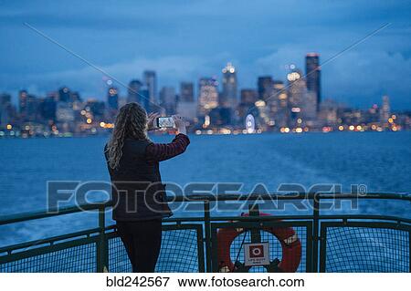 Stock Photo - Caucasian woman on boat photographing urban waterfront with cell phone. Fotosearch