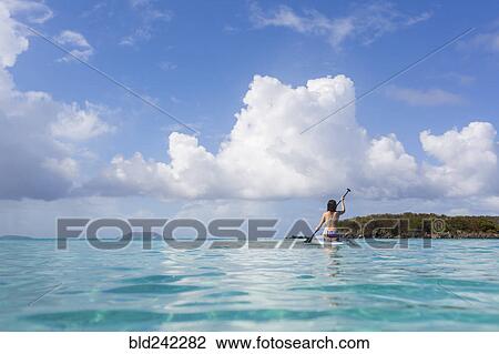 Stock Image - Caucasian woman paddling surfboard on tropical ocean. Fotosearch