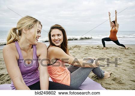 Stock Image - Caucasian woman performing yoga on beach near relaxing friends. Fotosearch
