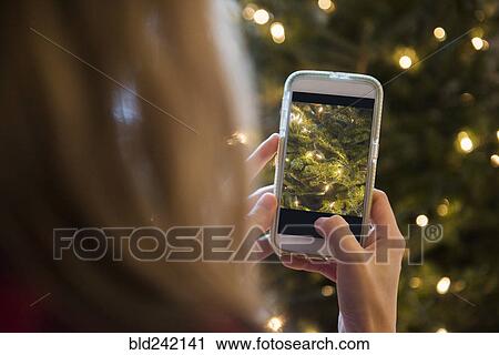 Stock Image - Caucasian woman photographing Christmas tree with cell phone. Fotosearch