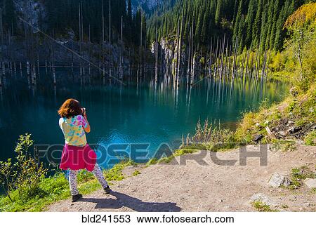 Stock Image - Caucasian woman photographing trees in lake. Fotosearch