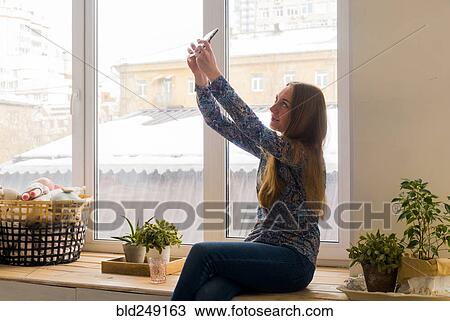 Stock Image - Caucasian woman sitting at window posing for cell phone selfie. Fotosearch