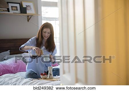 Stock Image - Caucasian woman sitting in bed pouring coffee. Fotosearch