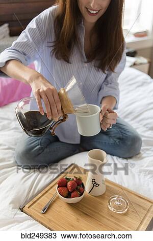 Stock Image - Caucasian woman sitting in bed pouring coffee. Fotosearch