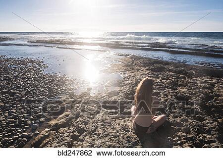 Stock Image - Caucasian woman sitting on beach wearing bikini. Fotosearch