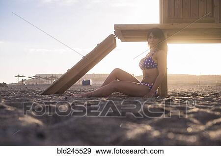 Stock Photo - Caucasian woman sitting on beach leaning on cabana. Fotosearch