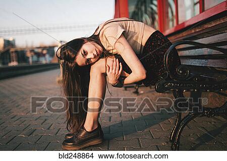 Picture - Caucasian woman sitting on bench holding legs. Fotosearch