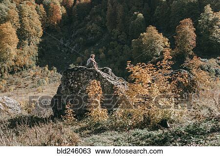 Stock Image - Caucasian woman sitting on boulder in autumn. Fotosearch