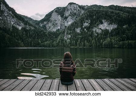 Stock Image - Caucasian woman sitting on dock admiring scenic view of mountain. Fotosearch