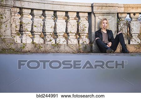 Stock Image - Caucasian woman sitting on ground leaning on wall. Fotosearch