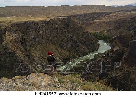 Stock Photo - Caucasian woman sitting on mountain rocks photographing river. Fotosearch