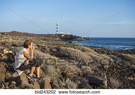 Stock Image - Caucasian woman sitting on rocks at ocean. Fotosearch