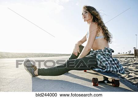 Stock Image - Caucasian woman sitting on skateboard on path at beach. Fotosearch