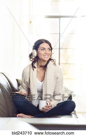 Stock Image - Caucasian woman sitting on sofa listening to headphones. Fotosearch