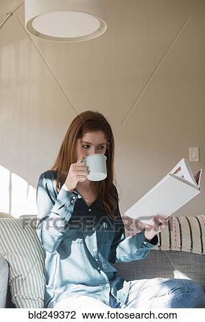 Stock Image - Caucasian woman sitting on sofa drinking coffee and reading book. Fotosearch