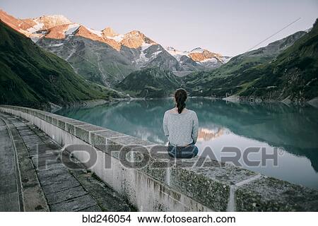 Picture - Caucasian woman sitting on wall at mountain lake. Fotosearch