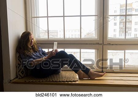 Stock Image - Caucasian woman sitting on window sill reading digital tablet. Fotosearch