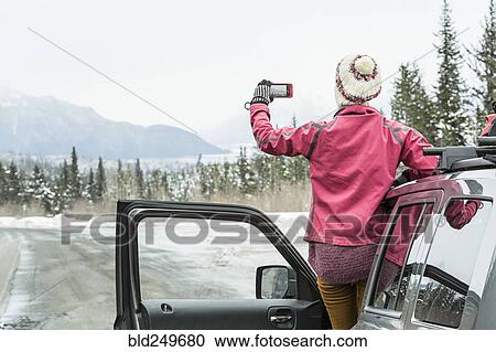 Stock Image - Caucasian woman standing in car in winter photographing scenic view. Fotosearch