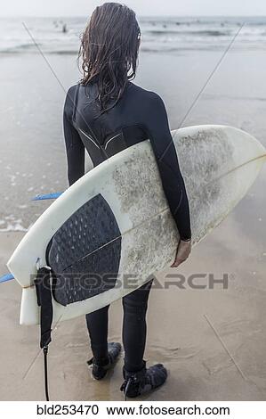 Stock Image - Caucasian woman standing on beach holding surfboard. Fotosearch