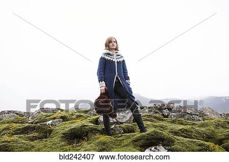 Stock Photo - Caucasian woman standing on mossy rocks holding fur hat. Fotosearch