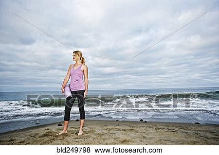 Stock Photo - Caucasian woman standing on ocean beach holding exercise mat. Fotosearch