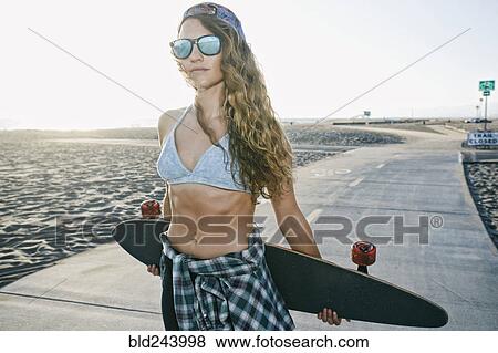 Stock Photo - Caucasian woman standing on path at beach with skateboard. Fotosearch