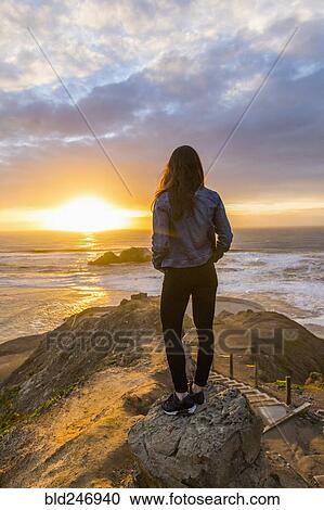 Stock Image - Caucasian woman standing on rock near ocean at sunset. Fotosearch