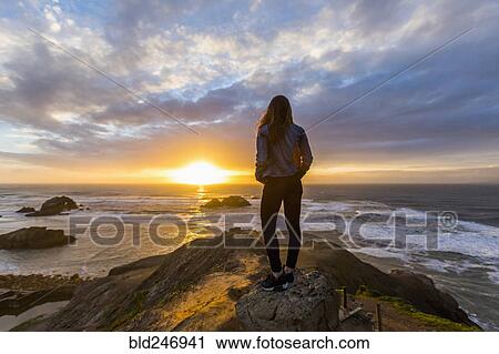 Stock Image - Caucasian woman standing on rock near ocean at sunset. Fotosearch