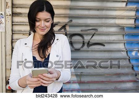 Stock Photo - Caucasian woman texting on cell phone near metal graffiti wall. Fotosearch