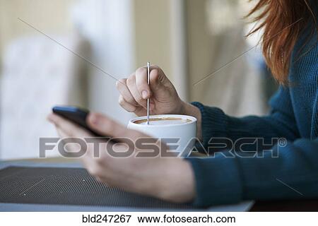 Stock Photo - Caucasian woman texting on cell phone and stirring coffee. Fotosearch