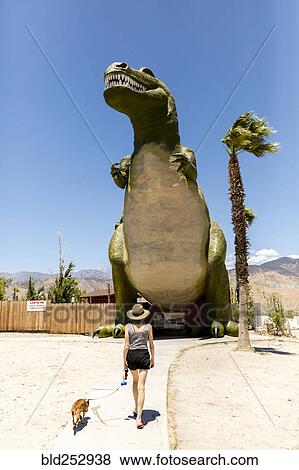 Stock Photo - Caucasian woman walking dog near giant dinosaur statue. Fotosearch