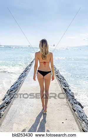 Stock Image - Caucasian woman wearing bikini walking on breakwall. Fotosearch