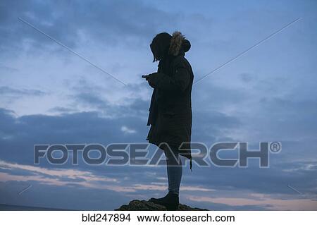 Picture - Caucasian woman wearing coat texting on cell phone at sunset. Fotosearch