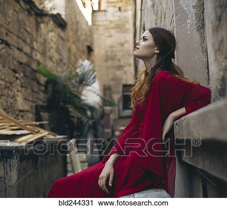 Stock Image - Caucasian woman wearing red dress sitting on bench in alley. Fotosearch