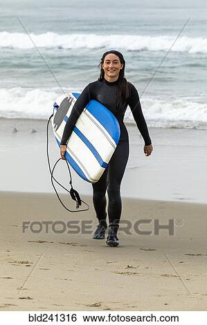 Stock Photograph - Caucasian woman wearing wetsuit carrying surfboard on beach. Fotosearch
