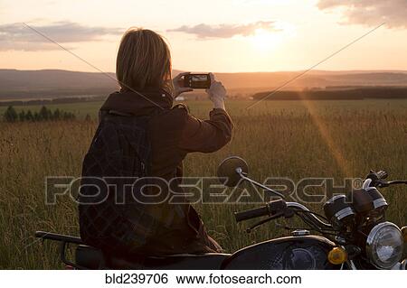 Caucasian woman with motorcycle in field photographing sunset View Large Photo Image Stock Photograph - Caucasian woman with motorcycle in field photographing sunset. Fotosearch
