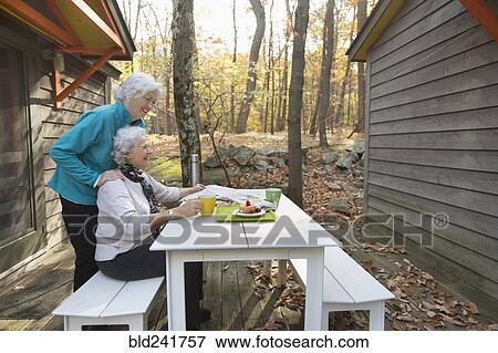 Stock Photo - Caucasian women enjoying breakfast outdoors near cabin. Fotosearch