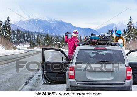 Stock Image - Caucasian women photographing from car with snowboards. Fotosearch