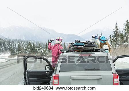Picture - Caucasian women standing in car in winter photographing scenic view. Fotosearch