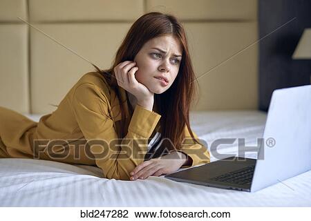 Stock Image - Confused Caucasian woman laying on bed using laptop. Fotosearch