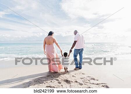 Stock Photo - Couple holding hands of baby daughter walking on beach. Fotosearch