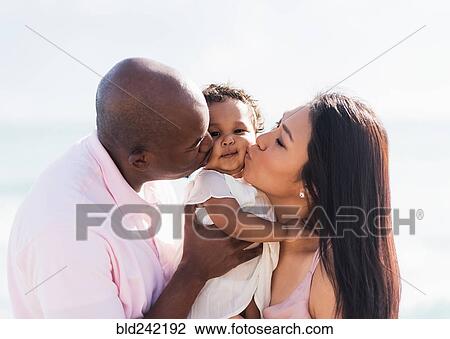 Stock Image - Couple kissing cheeks of baby daughter. Fotosearch