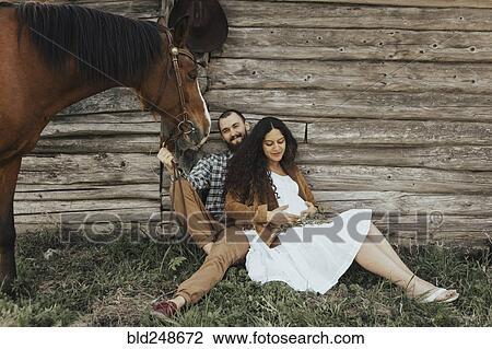Stock Image - Couple sitting in grass near barn with horse. Fotosearch