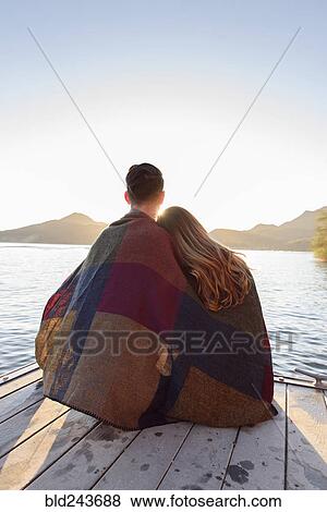 Stock Photo - Couple sitting on dock at river wrapped in blanket. Fotosearch