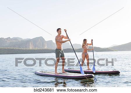 Stock Image - Couple standing on paddleboards in river. Fotosearch