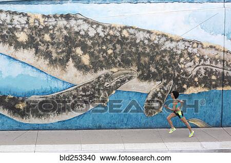 Stock Image - Distant Mixed Race woman running on sidewalk near mural. Fotosearch