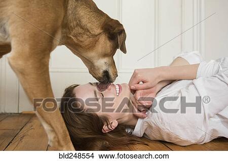 Picture - Dog licking nose of Caucasian woman laying on floor. Fotosearch