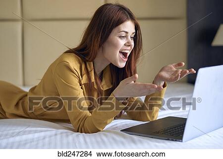Picture - Excited Caucasian woman laying on bed using laptop. Fotosearch