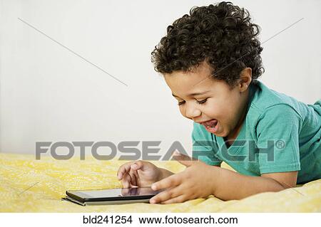 Excited Mixed Race boy laying on bed using digital tablet View Large Photo Image Picture - Excited Mixed Race boy laying on bed using digital tablet. Fotosearch