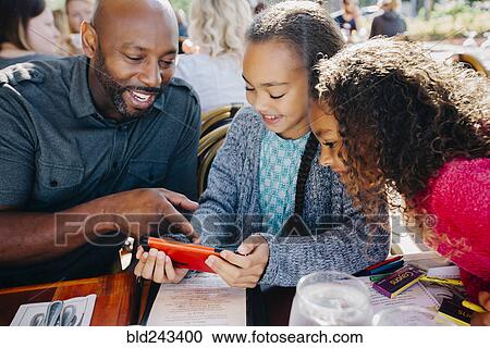 Stock Image - Father ad daughters texting on cell phone at restaurant. Fotosearch